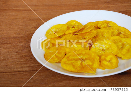 Dried and fried banana slices place on white plate isolated on wood texture background. This is local Malaysian traditional junk food call Kerepek Pisang. Dried and fried banana slices place on white plate isolated on wood texture background. This is local Malaysian traditional junk food call Kerepek Pisang. 78937313