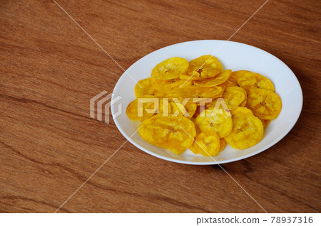 Dried and fried banana slices place on white plate isolated on wood texture background. This is local Malaysian traditional junk food call Kerepek Pisang. Dried and fried banana slices place on white plate isolated on wood texture background. This is local Malaysian traditional junk food call Kerepek Pisang. 78937316
