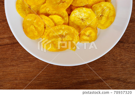 Dried and fried banana slices place on white plate isolated on wood texture background. This is local Malaysian traditional junk food call Kerepek Pisang. Dried and fried banana slices place on white plate isolated on wood texture background. This is local Malaysian traditional junk food call Kerepek Pisang. 78937320