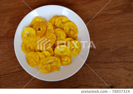 Dried and fried banana slices place on white plate isolated on wood texture background. This is local Malaysian traditional junk food call Kerepek Pisang. Dried and fried banana slices place on white plate isolated on wood texture background. This is local Malaysian traditional junk food call Kerepek Pisang. 78937324