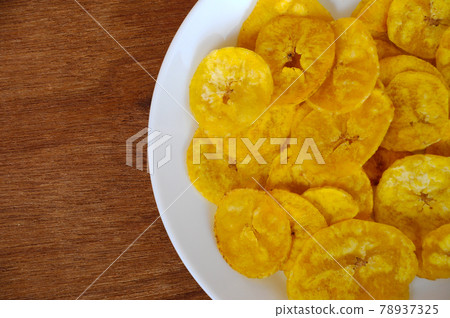 Dried and fried banana slices place on white plate isolated on wood texture background. This is local Malaysian traditional junk food call Kerepek Pisang. Dried and fried banana slices place on white plate isolated on wood texture background. This is local Malaysian traditional junk food call Kerepek Pisang. 78937325