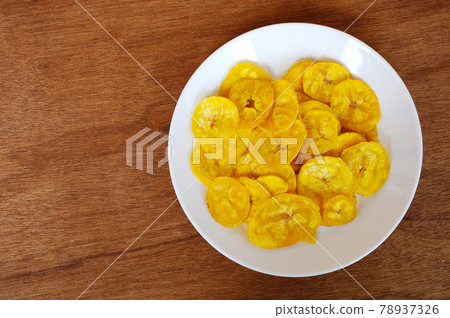 Dried and fried banana slices place on white plate isolated on wood texture background. This is local Malaysian traditional junk food call Kerepek Pisang. Dried and fried banana slices place on white plate isolated on wood texture background. This is local Malaysian traditional junk food call Kerepek Pisang. 78937326