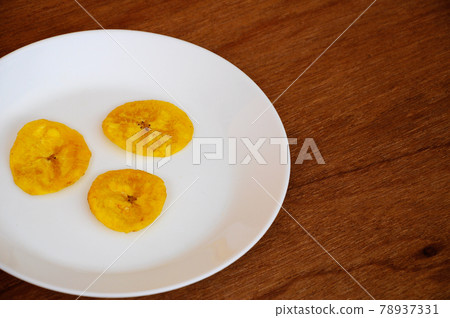 Dried and fried banana slices place on white plate isolated on wood texture background. This is local Malaysian traditional junk food call Kerepek Pisang. Dried and fried banana slices place on white plate isolated on wood texture background. This is local Malaysian traditional junk food call Kerepek Pisang. 78937331