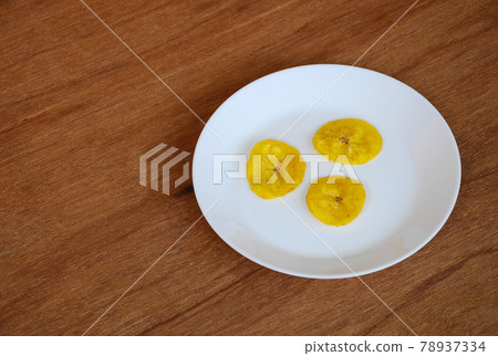 Dried and fried banana slices place on white plate isolated on wood texture background. This is local Malaysian traditional junk food call Kerepek Pisang. Dried and fried banana slices place on white plate isolated on wood texture background. This is local Malaysian traditional junk food call Kerepek Pisang. 78937334