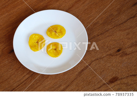 Dried and fried banana slices place on white plate isolated on wood texture background. This is local Malaysian traditional junk food call Kerepek Pisang. Dried and fried banana slices place on white plate isolated on wood texture background. This is local Malaysian traditional junk food call Kerepek Pisang. 78937335