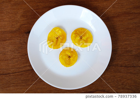 Dried and fried banana slices place on white plate isolated on wood texture background. This is local Malaysian traditional junk food call Kerepek Pisang. Dried and fried banana slices place on white plate isolated on wood texture background. This is local Malaysian traditional junk food call Kerepek Pisang. 78937336