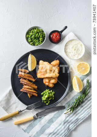British Traditional Fish and chips with minty mashed peas detail and a slice of lemon. on black round plate over white lintn and stone surface top view British Traditional Fish and chips with minty mashed peas detail and a slice of lemon. on black round plate over white lintn and stone surface top view 78941407
