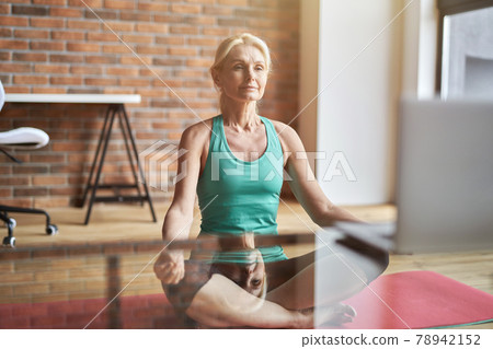 Relaxed mature blonde woman sitting in lotus position on the floor and watching online video training at home in the daytime Relaxed mature blonde woman sitting in lotus position on the floor and watching online video training at home in the daytime 78942152