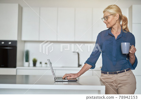 Attractive blonde businesswoman in glasses having a cup of coffee and using laptop in the morning, standing in the kitchen while working from home Attractive blonde businesswoman in glasses having a cup of coffee and using laptop in the morning, standing in the kitchen while working from home 78942153