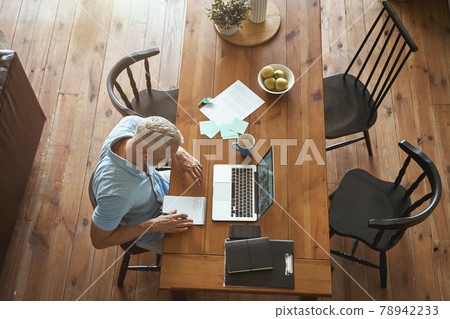Top view of a mature caucasian businessman making some notes and using laptop while sitting at table in his modern apartment Top view of a mature caucasian businessman making some notes and using laptop while sitting at table in his modern apartment 78942233