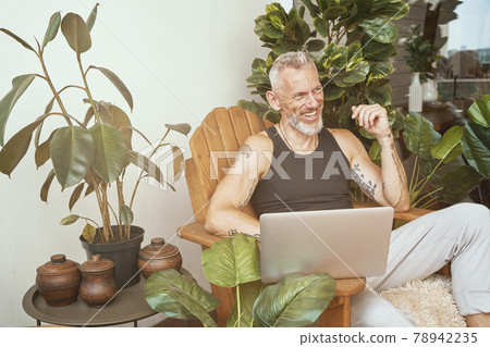 Happy middle aged caucasian man in casual clothes using laptop while sitting on the balcony or terrace of his modern apartment Happy middle aged caucasian man in casual clothes using laptop while sitting on the balcony or terrace of his modern apartment 78942235