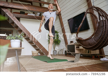 Sport during self isolation and covid 19 outbreak. Active middle aged man exercising in the living room of his modern apartment while watching online workout on laptop 78942286