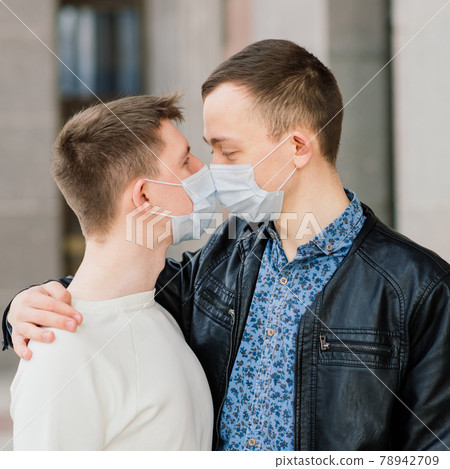 Young gay couple wearing medical mask, hugging and kissing at the city. Young gay couple wearing medical mask, hugging and kissing at the city. 78942709
