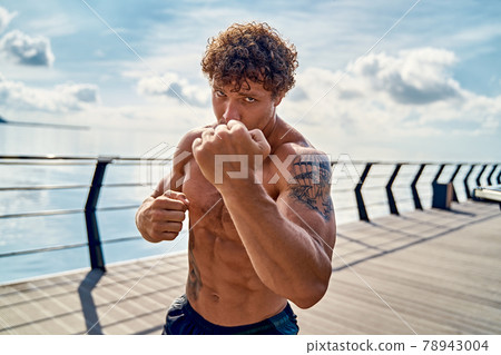 Muscular young man athlete standing and practicing shadow boxing outdoors early in the morning on pier by the sea Muscular young man athlete standing and practicing shadow boxing outdoors early in the morning on pier by the sea 78943004