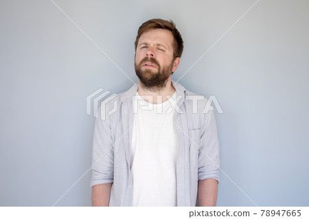 Caucasian, bearded man in casual clothes looks suspicious while standing against gray background. 78947665