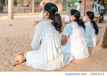Buddhist Female devotees worshipping at Jaya Sri Maha Bhodi while sitting on the sandy soil in white dresses. Concept of new normal, wearing a face mask while chanting prayers. 78954513