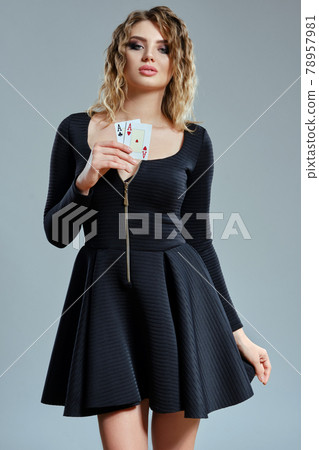Blonde maiden with curly hair, in black short dress showing two aces, posing against gray background. Gambling entertainment, poker, casino. Close-up. Blonde maiden with curly hair, in black short dress showing two aces, posing against gray background. Gambling entertainment, poker, casino. Close-up. 78957981