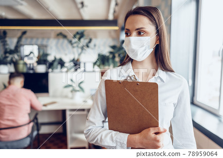 Young businesswoman in face mask standing in office and holding clipboard with documents 78959064