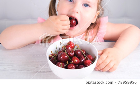 Beautiful cute little girl sitting at a light wooden table with a plate of cherries and eating a Beautiful cute little girl sitting at a light wooden table with a plate of cherries and eating a 78959956