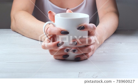 Female hands are holding a white cup of coffee on a light wooden table. The concept of suffering Female hands are holding a white cup of coffee on a light wooden table. The concept of suffering 78960027