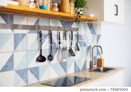 Kitchen spatulas handing on railing on a tiled wall with blue geometric pattern 78965004
