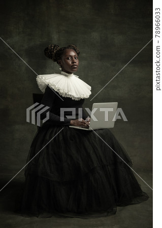 Portrait of medieval African young woman in black vintage dress with big white collar posing isolated on dark green background. Using laptop 78966033