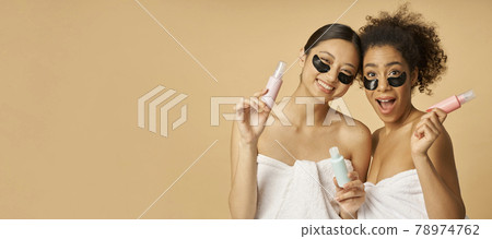 Website header of Two cute, young women with hydro gel under eye patches looking excited, holding bottles with beauty product, posing isolated over beige background 78974762