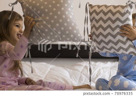 home, comfort, childhood, care, friendship - two little funny happy authentic baby toddler sibling kids children sit on white bed look at camera play fight with pillows soft cozy sleepy mood indoors 78976335