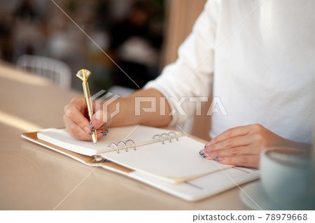 Well groomed woman hand holding gold pen and writing notes with gold pen in notebook beside window. 78979668