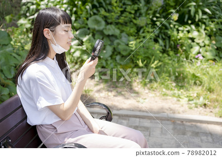 一個女孩坐在公園的長椅上,戴著下巴面罩,用手持風扇冷卻 一個女孩坐在公園的長椅上,戴著下巴面罩,用手持風扇冷卻 78982012