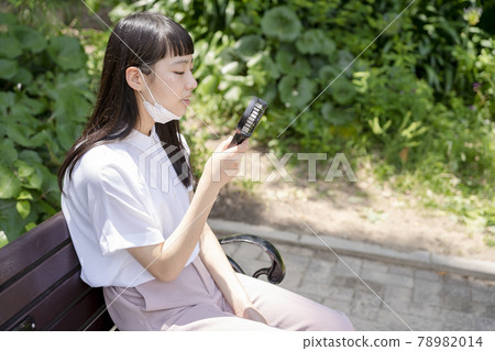 一個女孩坐在公園的長椅上,戴著下巴面罩,用手持風扇冷卻 一個女孩坐在公園的長椅上,戴著下巴面罩,用手持風扇冷卻 78982014