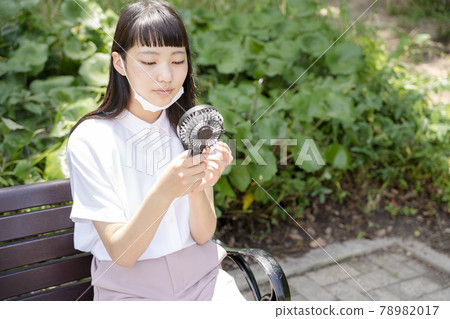 A girl sitting on a park bench, wearing a chin mask and cooling with a handheld fan A girl sitting on a park bench, wearing a chin mask and cooling with a handheld fan 78982017