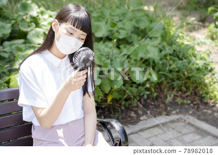 A girl sitting on a park bench, wearing a mask and cooling with a handheld fan A girl sitting on a park bench, wearing a mask and cooling with a handheld fan 78982018