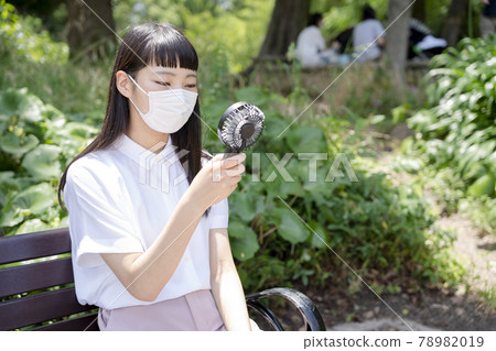 A girl sitting on a park bench, wearing a mask and cooling with a handheld fan A girl sitting on a park bench, wearing a mask and cooling with a handheld fan 78982019