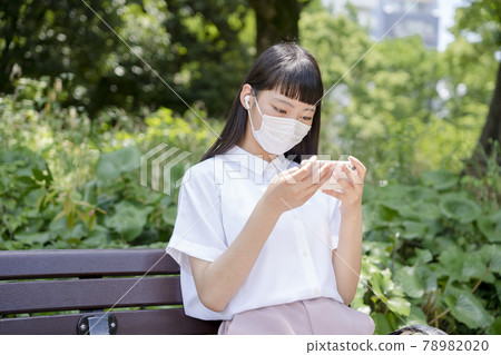 A girl sitting on a park bench, wearing a mask and watching a video on her smartphone A girl sitting on a park bench, wearing a mask and watching a video on her smartphone 78982020