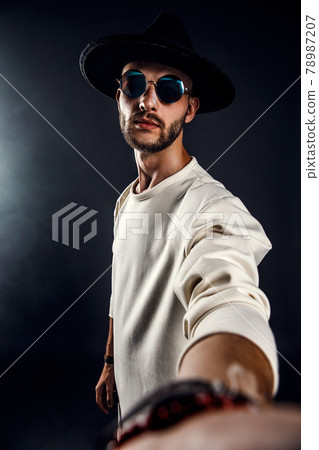 Portrait of a man in a wide-brimmed hat and sunglasses looking at camera while taking selfie on dark background. Portrait of a man in a wide-brimmed hat and sunglasses looking at camera while taking selfie on dark background. 78987207
