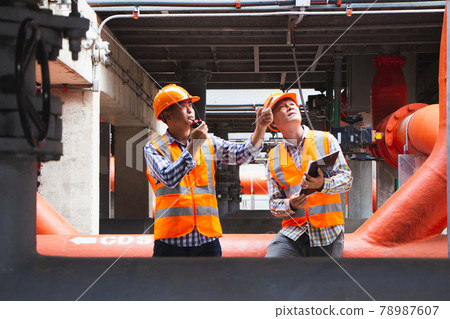 Asian male engineer inspecting pipe fittings and chillers, refrigeration plant refrigeration plant in  factory. men mechanics wear helmets and Reflective Safety Vests, use laptops, notebooks, and 78987607