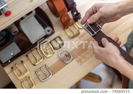 Closeup hands of professional tanner choosing belt buckle on wooden table at leather workshop 78989555
