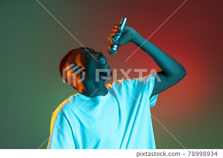 Young african man singing with microphone on neon gradient background. Concept of emotion, action, facial expression. Young african man singing with microphone on neon gradient background. Concept of emotion, action, facial expression. 78998934