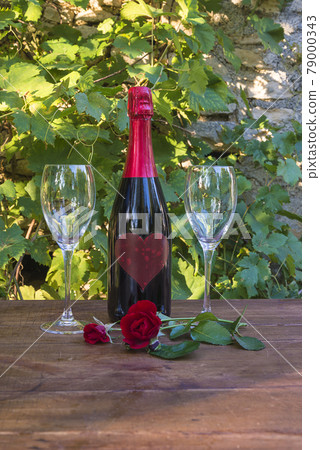 Bottle of red champagne, two wineglasses and rose flowers on wooden table in front of grapes leaves; vertical picture Bottle of red champagne, two wineglasses and rose flowers on wooden table in front of grapes leaves; vertical picture 79000343
