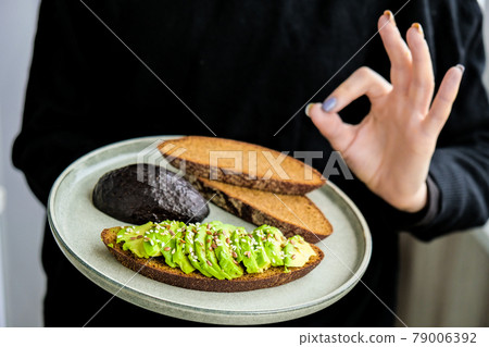 Female hand holding plate with Ingredients for healthy avocado toast. ripe hass avocado, wholegrain bread, sesame flax seeds. Vegan keto diet. Healthy lifestyle. 79006392