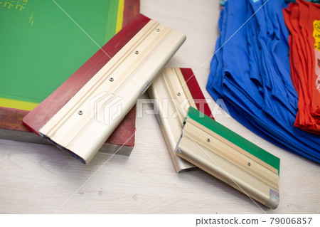 Serigraphy silk screen print process at clothes factory. Frame template stencil, squeegee and plastisol paint at table with cloth Serigraphy silk screen print process at clothes factory. Frame template stencil, squeegee and plastisol paint at table with cloth 79006857