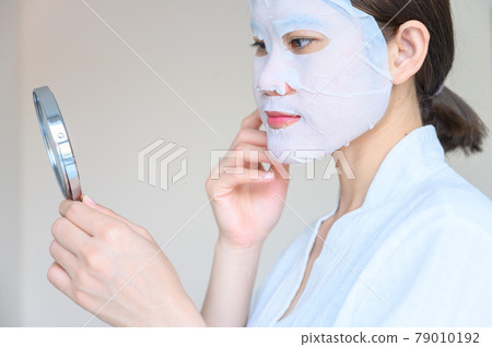 Portrait of young woman looking her face in mirror after applying facial mask for enhance her skin. Facial mask is a creamy or thick pasted mask applied to clean or smoothen the face. 79010192