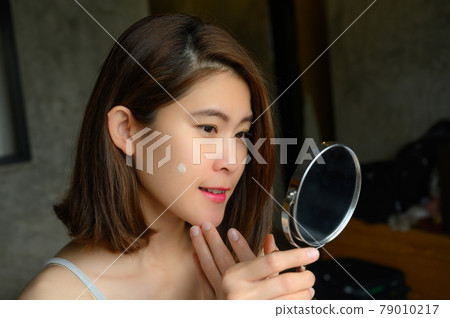 Portrait of young Asian woman holding mirror and applying acne cream/moisturizer on her face for treat her skin. Conceptual of beauty and cosmetic. Portrait of young Asian woman holding mirror and applying acne cream/moisturizer on her face for treat her skin. Conceptual of beauty and cosmetic. 79010217