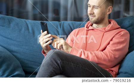 A man uses a smartphone on his home couch while relaxing at home. Applications, calls, internet concept. A man uses a smartphone on his home couch while relaxing at home. Applications, calls, internet concept. 79019216