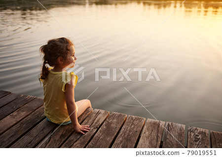 An adorable baby girl sits on a wooden pier by the lake and enjoys the warm of a summer evening. The beautiful light of the sunset is reflected in the water of the lake. An adorable baby girl sits on a wooden pier by the lake and enjoys the warm of a summer evening. The beautiful light of the sunset is reflected in the water of the lake. 79019551