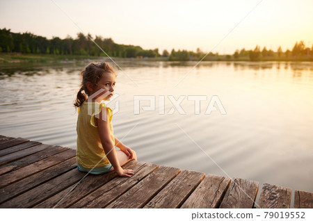 Cute baby girl relaxing on the pier at sunset. Warm summer evening. Heat summer, summer evenings in nature, spending time in the fresh air Cute baby girl relaxing on the pier at sunset. Warm summer evening. Heat summer, summer evenings in nature, spending time in the fresh air 79019552