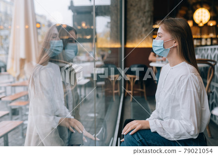 Two women in protective masks opposite each other, window between them. Two women in protective masks opposite each other, window between them. 79021054