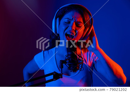 Caucasian woman in headphones singing into a microphone in neon light on a black background. An emotional girl is recording a song in a recording studio Caucasian woman in headphones singing into a microphone in neon light on a black background. An emotional girl is recording a song in a recording studio 79021202