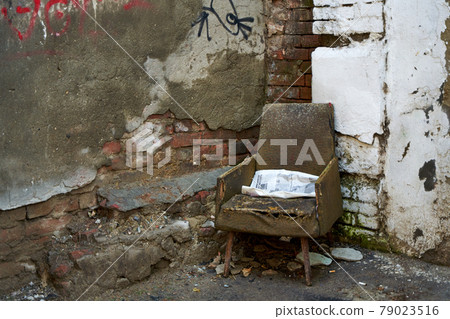 The atmosphere of devastation. An old decrepit chair with a snowy newspaper stands on the street next to the crumbling wall of an old building The atmosphere of devastation. An old decrepit chair with a snowy newspaper stands on the street next to the crumbling wall of an old building 79023516